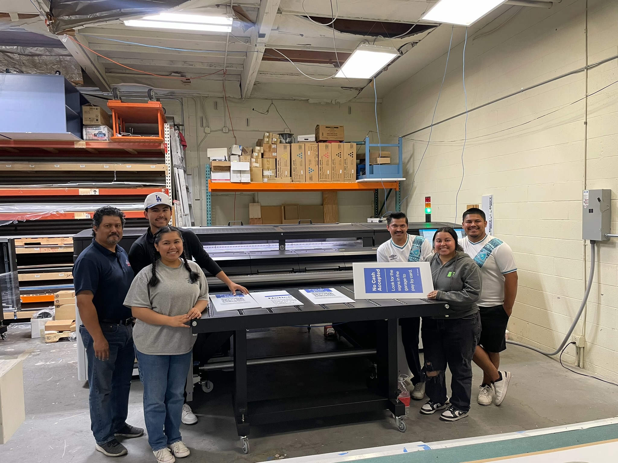 Six members of the Accurate DPS team gathered around a large-format printer with completed parking signs in the workshop