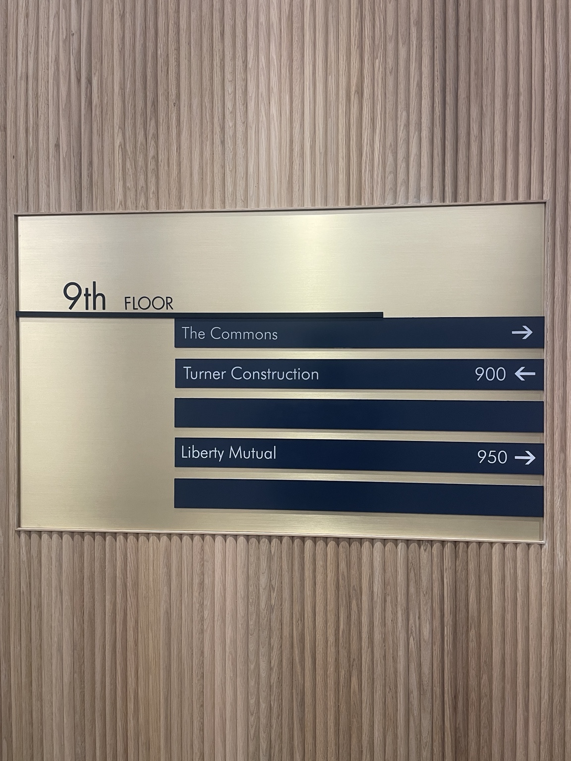 Gold-colored 9th-floor directory with blue tenant-name panels and directional arrows on a wood wall