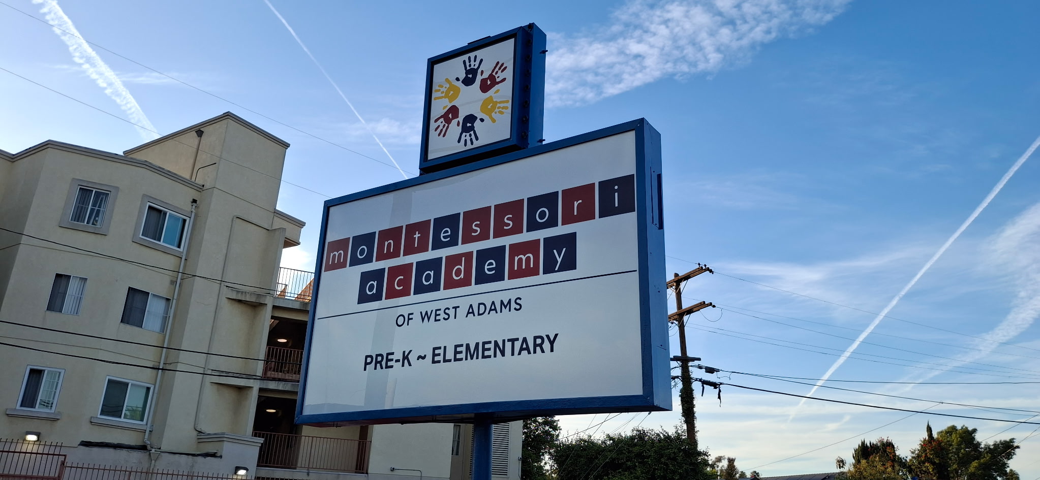 Blue-framed exterior sign for Montessori Academy of West Adams, with a handprint graphic above the school name