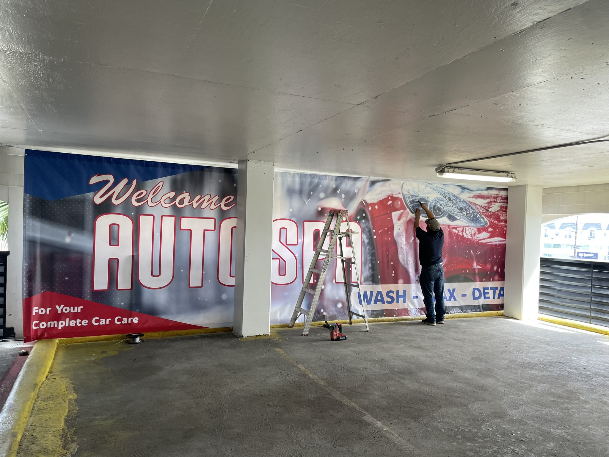 An installer applying a large wall graphic beside a ladder in a covered parking area